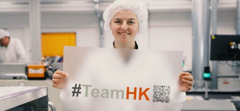 Hoffmann + Krippner - Mitarbeiterin hebt bedruckte Folie mit #TeamHK in die Kamera